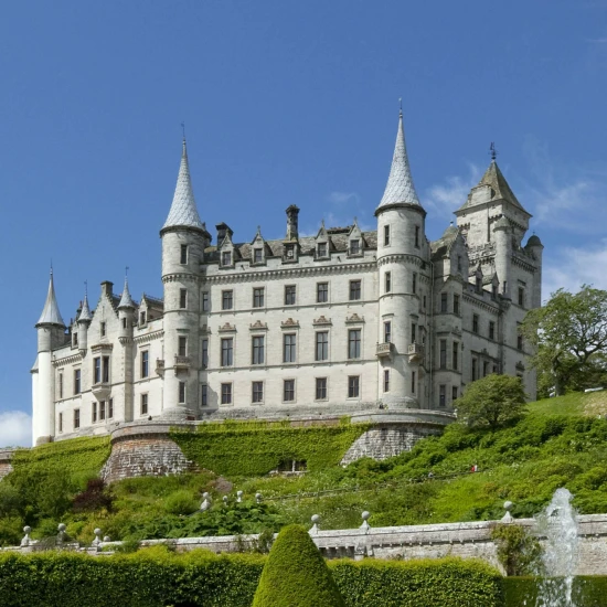 A stunning castle with multiple spires, surrounded by lush greenery and vibrant gardens under a clear blue sky.