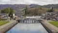 A scenic view of a canal with a lock, surrounded by charming buildings, trees, and hills under a cloudy sky.