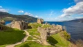 Ruins of a castle near a lake, surrounded by green hills under a blue sky with scattered clouds. Ideal for exploring history.