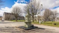 A historic monument stands on a roundabout, surrounded by bare trees and buildings under a blue sky with clouds.