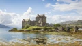 A scenic view of a historic castle surrounded by water and lush landscapes under a bright, partly cloudy sky.