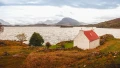A scenic view of a white cottage with a red roof by a lake, surrounded by mountains and autumnal landscape.