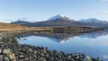Scenic view of snow-capped mountains reflected in calm water, surrounded by rocky shoreline and golden grassland under clear skies.