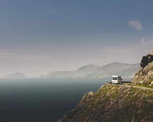A tour bus driving along a scenic coastal road with mountains and ocean in the background, showcasing UK's natural beauty.
