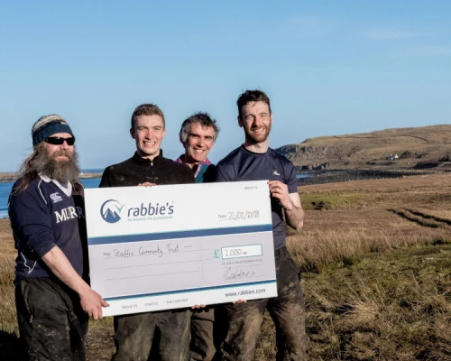 Group holding a large check for £2,000 outdoors, with scenic hills and coast in the background, promoting community support.