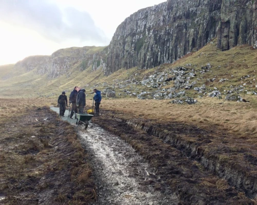 Group of people working on a muddy trail in a scenic landscape, with hills and rocky cliffs in the background.