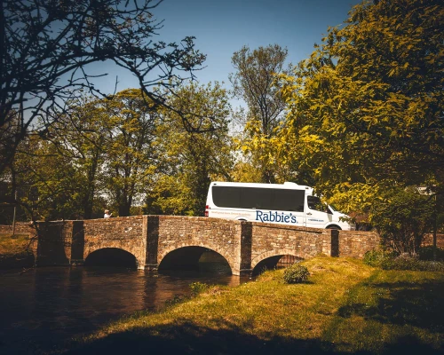 A Rabbie's Tours van crosses a picturesque stone bridge surrounded by greenery and trees, reflecting a serene UK landscape.