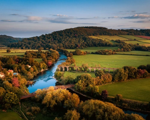 Scenic aerial view of lush green hills, a winding river, and an old stone arch bridge amidst colorful autumn foliage.