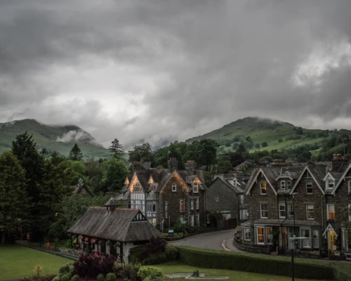 A quaint village with stone houses under a dramatic cloudy sky, surrounded by lush green hills.
