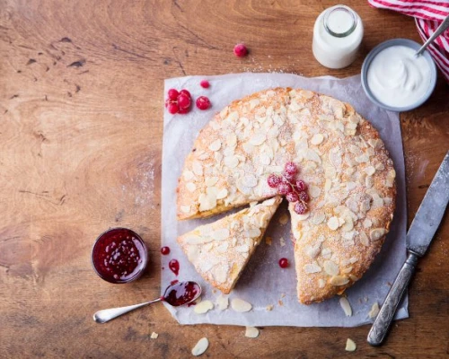 A freshly baked almond cake topped with raspberries, served with jam and cream on a rustic wooden table.