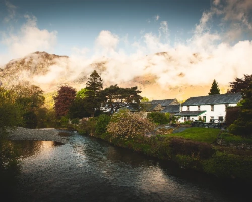 Scenic view of a quaint cottage by a river, surrounded by lush greenery and misty mountains in the background.