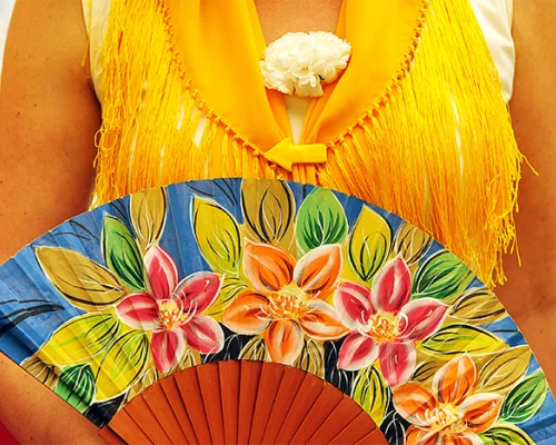 A person wearing a bright yellow fringed shawl holds a colorful hand-painted fan adorned with floral patterns.