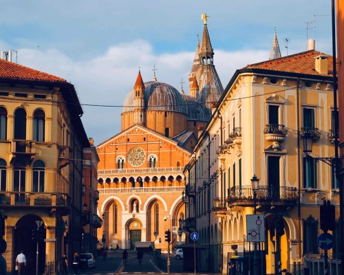Historic buildings with domes and towers flanking a church, bathed in warm sunlight, evoke a charming urban atmosphere.