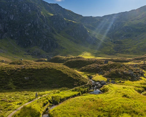 A serene landscape of rolling green hills and a winding path, illuminated by sunlight streaming through the mountains.