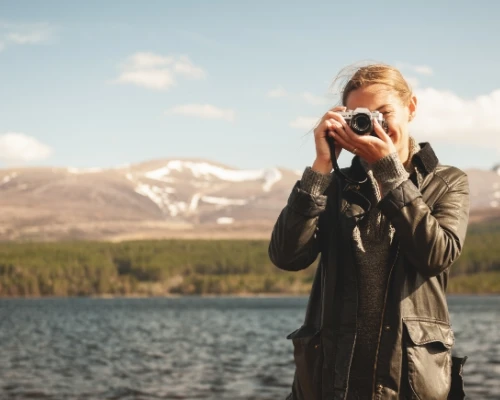 A person with a camera captures the scenic beauty of a lake surrounded by mountains under a clear blue sky.