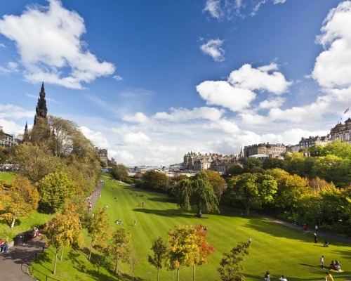 A vibrant park in Edinburgh with green lawns, colorful trees, and a backdrop of historical buildings under a bright blue sky.