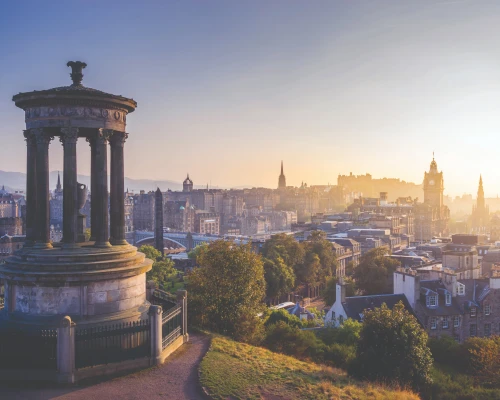 Sunrise over Edinburgh, featuring a historical monument with a panoramic view of the city skyline and soft morning light.