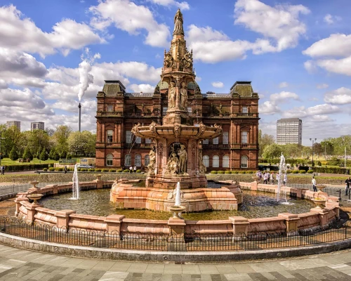 Victorian fountain in front of a grand building, surrounded by greenery and people, under a bright blue sky with fluffy clouds.