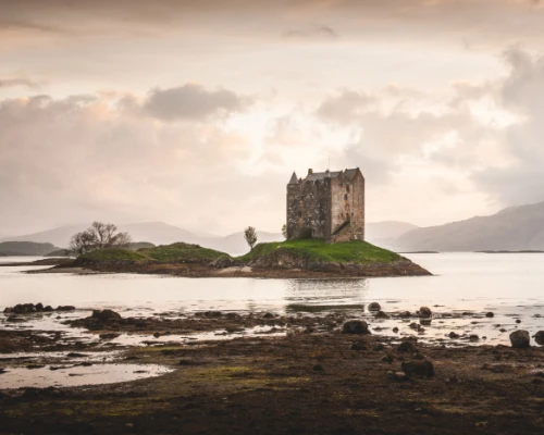 A solitary castle stands on a small island, surrounded by calm waters and distant mountains under a cloudy sky.