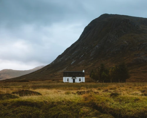 A solitary white house nestled in a grassy landscape beneath a dramatic mountain under overcast skies.