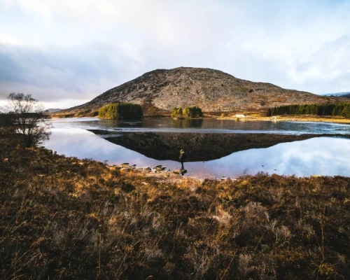 A serene landscape featuring a lake reflecting a mountain, with a lone figure standing by the water's edge amidst nature.