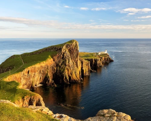 Stunning coastal view of a rocky cliff by the sea, featuring lush greenery and a distant lighthouse under a clear blue sky.