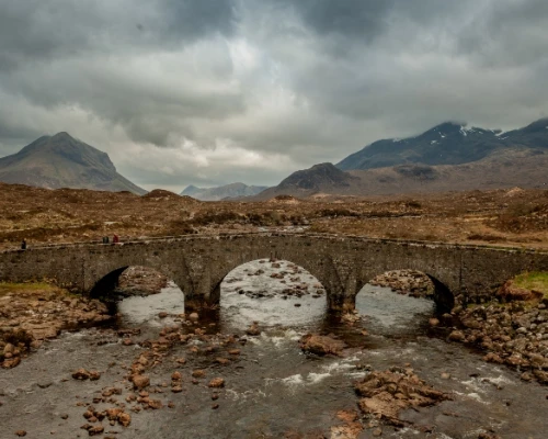 A stone bridge arches over a river, surrounded by rugged mountains under a cloudy sky in the UK.