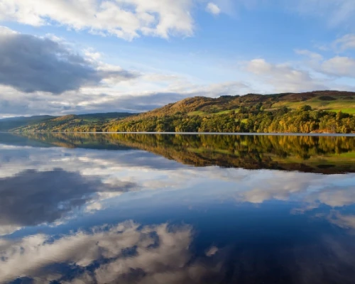 Scenic view of a calm lake reflecting lush green hills and a blue sky with fluffy clouds, showcasing the beauty of the UK.