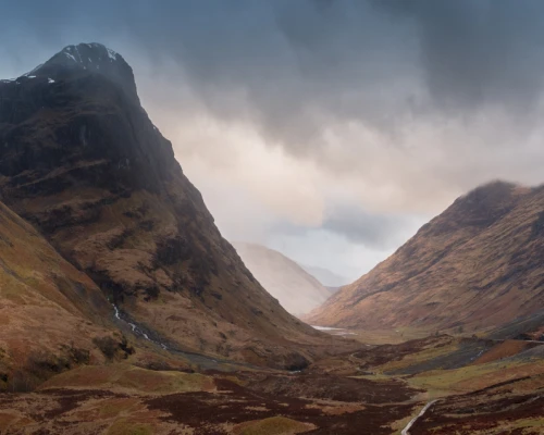 Dramatic Scottish landscape featuring towering mountains and a winding valley shrouded in moody clouds. Perfect for tours.