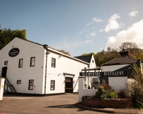 Glengoyne Distillery exterior featuring white buildings, black signage, and a clear blue sky, surrounded by greenery.