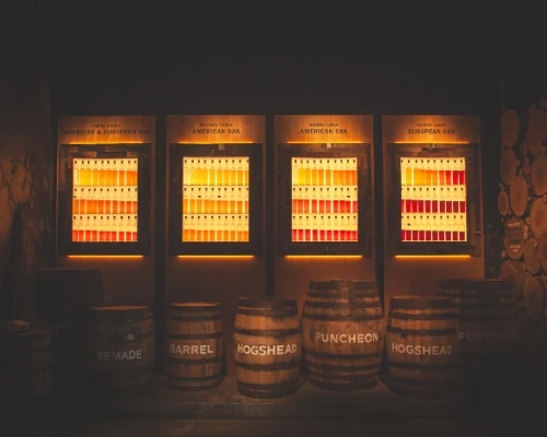 Three illuminated display boards showcase whiskey aging options above wooden barrels labeled with various types.