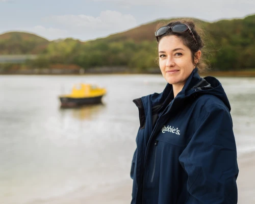 A woman in a Rabbie's jacket stands on a beach, with a yellow boat in the background and hills visible behind her.
