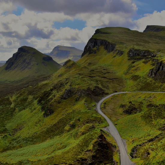 Breathtaking view of lush green hills and winding roads, with a serene lake and dramatic cliffs under a partly cloudy sky.
