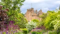 A lush garden with vibrant flowers frames a historic castle under a partly cloudy sky.
