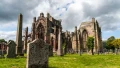 Ruins of a Gothic church surrounded by gravestones and lush greenery under a cloudy sky, hinting at rich history.
